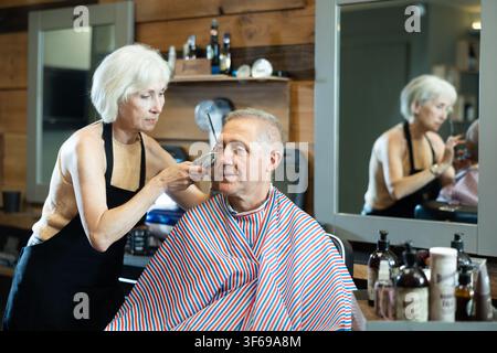 Le maître expérimenté de la femme mature fait effectue la coupe de cheveux pour l'homme âgé avec tondeuse Banque D'Images