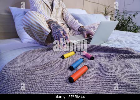 Jeune homme beau en pyjama avec différentes cigarettes électroniques jetables et ordinateur portable sur le lit à la maison Banque D'Images