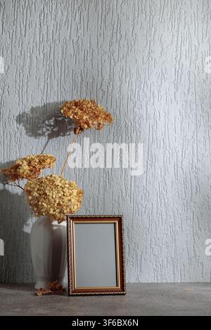 Cadre photo vide et vase blanc avec des fleurs sèches sur un fond de mur gris. Cadre en bois avec ornement doré. Placez votre image dans le cadre. Co Banque D'Images