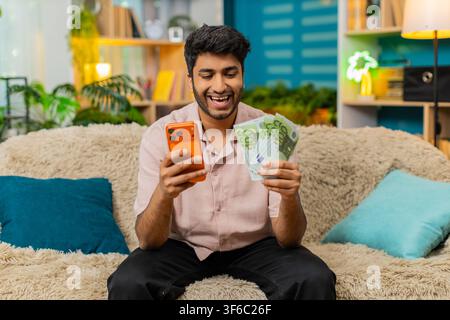 Homme indien sur un canapé à la maison tient des billets en euros avec un smartphone et sourit après une victoire d'application de jeu de casino ou de paris sportifs. Jeune arabe sur un canapé à la maison célèbre son profit avec un visage excité dans le salon Banque D'Images