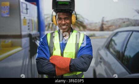 Homme portant un casque de soudure et gilet réfléchissant, les bras croisés et souriant dans la rue près d'une pompe de voiture ; sécurité de confiance. Banque D'Images