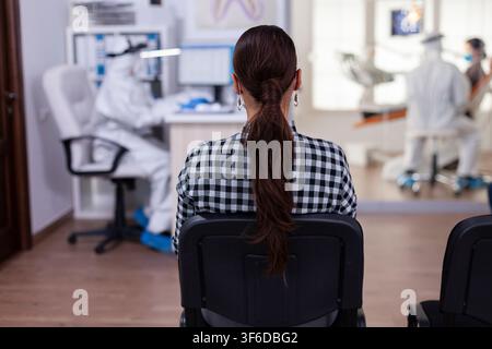 En réception, un dentiste portant des équipements de protection parle avec une patiente tandis que des soins dentaires de routine se poursuivent à proximité dans la clinique. Banque D'Images