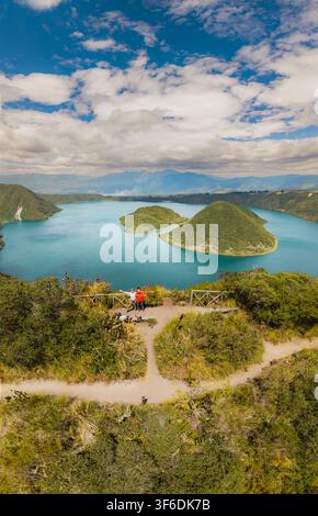 Vue aérienne de la Laguna de Cuicocha avec son lac de cratère turquoise, ses îles et ses pentes verdoyantes sous un ciel bleu nuageux, avec deux personnes debout sur Banque D'Images
