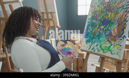 Femme peintre, brune hispanique, tient une palette de peinture en bois et un pinceau tout en souriant et en appliquant des traits colorés sur une toile dans un studio d'art ; cre Banque D'Images