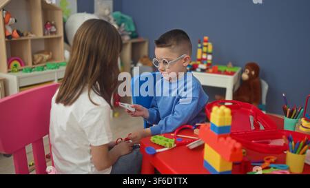 Une fille et un garçon jouent au docteur, le garçon place un stéthoscope jouet sur le bras de la fille dans une salle de jeux dans un bâtiment ; jeu compatissant. Banque D'Images