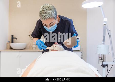 Esthéticienne professionnelle portant un masque de protection et des gants applique un masque facial à une cliente dans une clinique de beauté moderne lors d'un soin relaxant Banque D'Images