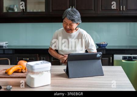 Une femme asiatique âgée souriante utilise une tablette sur un plan de travail de cuisine, profitant d'une conversation en ligne ou d'une recherche de recettes à côté de fruits frais et d'ingrédients de garde-manger Banque D'Images