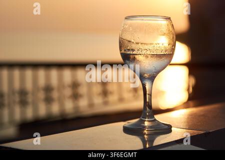 Close-up d'un verre refroidi d'ouzo grec traditionnel avec des gouttes de condensation à la surface, posé sur un balcon surplombant la mer au coucher du soleil Banque D'Images