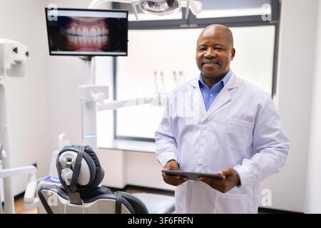 Dentiste masculin noir tenant une tablette numérique à côté du fauteuil dentaire dans une clinique moderne Banque D'Images