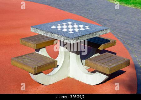 Table d'échecs en béton fixe d'extérieur avec plateau en damier carrelé et quatre sièges en bois. La table de jeu du parc public est posée sur un revêtement de sol synthétique rouge Banque D'Images