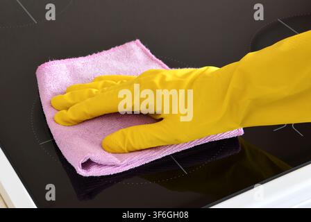 Main gantée de jaune nettoyant une plaque à induction avec un chiffon en microfibres rose Banque D'Images