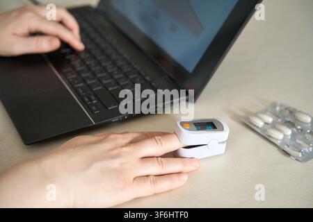 Oxymètre de pouls à placement sur le doigt affichant la lecture à côté d’un ordinateur portable et d’un emballage sous blister sur le bureau pendant le travail à distance. Banque D'Images