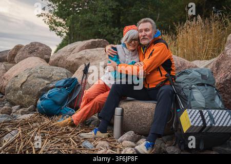 Aventure touristique de randonnée. Couple de seniors, homme et femme, voyageant ensemble et profitant des loisirs en plein air en randonnée, prenant une pause assis sur la plage. Personnes âgées heureuses, backpackers, randonneurs marchant en trekking dans la nature Banque D'Images