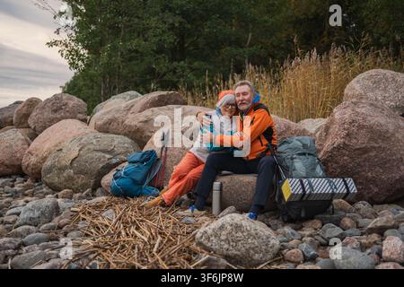 Aventure touristique de randonnée. Couple de seniors, homme et femme, voyageant ensemble et profitant des loisirs en plein air en randonnée, prenant une pause assis sur la plage. Personnes âgées heureuses, backpackers, randonneurs marchant en trekking dans la nature Banque D'Images