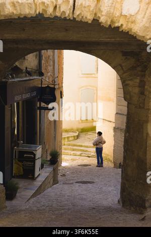 Vue à travers une arche ancienne en pierre vers une boulangerie et une touriste dans une ruelle étroite à Gordes, France. Banque D'Images