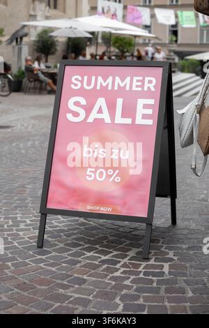 Villach, Autriche - 03 juillet 2026 : Panneau promotionnel pour une vente d'été avec des rabais jusqu'à 50 % dans une zone piétonne animée dotée de places de siège en plein air et de parasol Banque D'Images