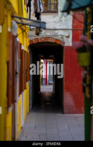 Burano, Italie – 20 juin 2026 : Perspective verticale regardant à travers une arche d’une ruelle étroite et sombre encadrée par des façades de bâtiments peintes en jaune vif et rouge Banque D'Images