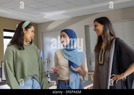 Diverses collègues féminines debout et discutant au bureau avec moniteur, ordinateurs portables, portant le hijab. Collègues, collaboration, réunion, dialogue, moderne, professionnel, amical Banque D'Images