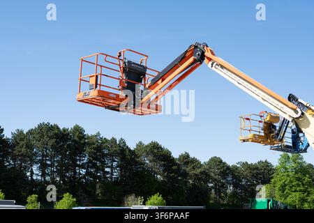 Plusieurs nacelles élévatrices, dont un modèle JLG orange, sont surélevées bien au-dessus du sol avec un fond d'arbres verts et d'un ciel bleu clair Banque D'Images