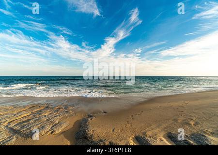 Le sable doré rencontre des vagues turquoise sous un ciel bleu éclatant. Des nuages clairsemés dérivent au-dessus, ajoutant de la douceur à l'horizon. Des empreintes de pas fanées laissent suggérer que Banque D'Images