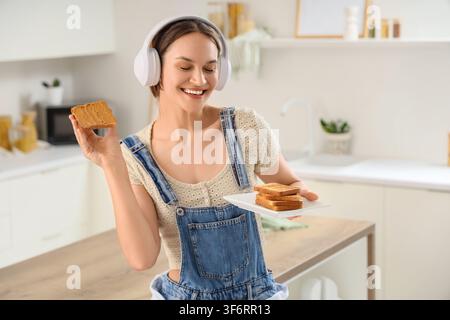 Jeune femme heureuse portant des écouteurs et une salopette en jean, tenant un toast au beurre de cacahuète et une assiette de toast nature dans une cuisine moderne Banque D'Images