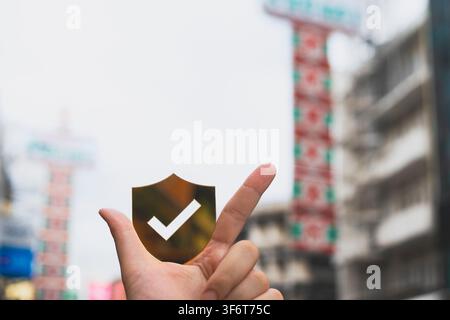 Main tenant une icône de bouclier doré avec une coche et un signe chinois flou dans un arrière-plan urbain, représentant l'assurance voyage, la sécurité à l'étranger. Banque D'Images