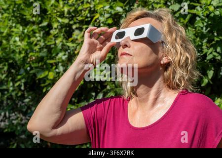 Femme blonde en rouge observe le soleil avec des lunettes de protection classiques pour éclipse solaire Banque D'Images