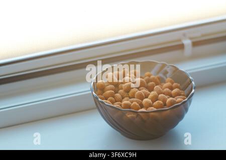 Vue rapprochée de cacahuètes grillées croustillantes dans un petit bol sombre posé sur un rebord de fenêtre lumineux avec une lumière naturelle filtrant à travers la vitre. Banque D'Images