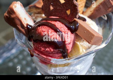 Close-up d'une tasse à sundae en verre remplie de boules de glace à la framboise, décorée de sirop de chocolat, de divers biscuits, de bâtonnets de gaufrettes et de barres de chocolat f Banque D'Images