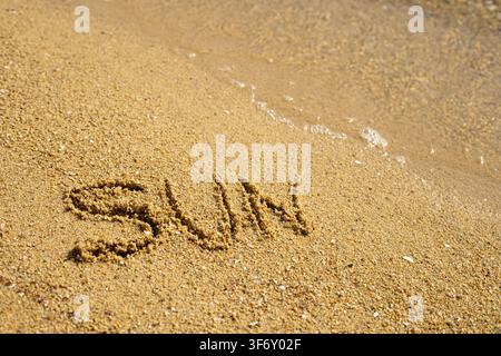 Gros plan du mot soleil écrit dans le sable au bord de l'eau. Mise au point nette sur les grains texturés avec de douces vagues de l'océan qui déferlent sur le rivage. Banque D'Images