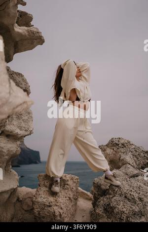 Woman Loungewear rocks jeune femme portant un pantalon de survêtement crème, debout sur des falaises côtières surplombant l'océan avec espace de copie Banque D'Images