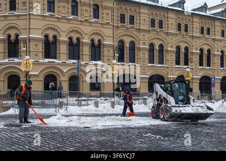 Moscou, Russie, 20 décembre 2025 Ouvriers municipaux en gilets de sécurité orange déblayant la neige avec des pelles et une chargeuse Bobcat sur le c Banque D'Images