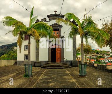 Jangão, Portugal - 06.12.2025 : Petite église blanche avec des accents de pierre volcanique sombre et des palmiers sur une place pavée à Madeira Banque D'Images