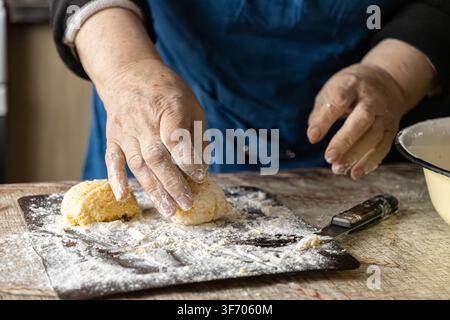 Vue rapprochée d'une femme âgée préparant la pâte pour des syrniki traditionnels sur une table en bois, mettant l'accent sur la préparation tactile de la nourriture faite maison et le temps Banque D'Images