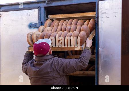 Un ouvrier en veste charge du pain fraîchement cuit sur les étagères coulissantes d'un camion, mettant en valeur un processus de boulangerie artisanal traditionnel, la logistique alimentaire et le da Banque D'Images