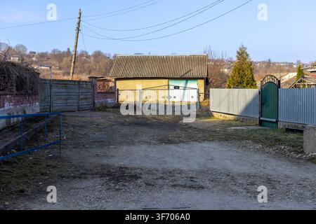 Un chemin de terre mène à une petite maison extérieure avec un toit en tôles ondulées et des clôtures dans un village tranquille, capturant l'atmosphère authentique de la paix r Banque D'Images