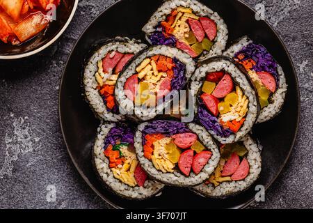 Une assiette noire remplie de kimbap (ou gimbap) aux garnitures colorées. Les ingrédients comprennent des saucisses, des cornichons, des œufs et des légumes. Banque D'Images