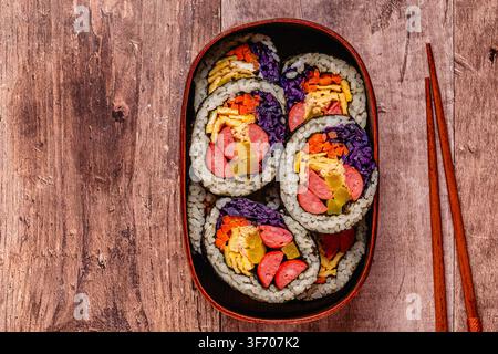 Kimbap (ou gimbap) coloré avec des légumes et de la saucisse dans une boîte en bois sur une surface en bois Banque D'Images