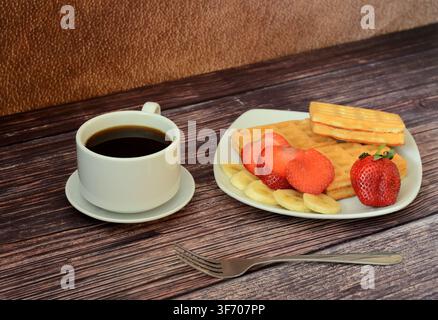 Une assiette de gaufres viennoises fraîches avec des fraises et des bananes sur une table en bois, à côté d'une tasse de café noir chaud sur une soucoupe. Gros plan. Banque D'Images