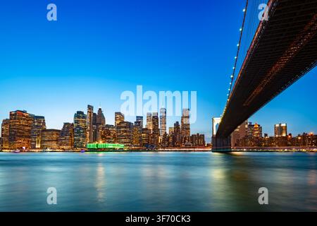 Vue spectaculaire du centre-ville de Manhattan avec l'emblématique pont de Brooklyn s'étendant sur l'eau alors que les lumières de la ville se reflètent sur la rivière. Banque D'Images