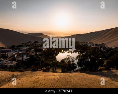 Vue aérienne de l'oasis de Huacachina, une oasis du désert entourée de dunes de sable massives, de palmiers et de bâtiments au coucher du soleil, Huacachina, Ica, Pérou. Banque D'Images