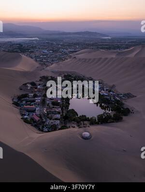 Vue aérienne de l'oasis de Huacachina entourée de dunes de sable imposantes et de palmiers au coucher du soleil à Huacachina, Ica, Pérou. Banque D'Images
