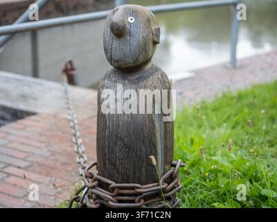 Figure de bollard en bois avec une chaîne enroulée autour, près d'une promenade riveraine sur une berge herbeuse. Banque D'Images