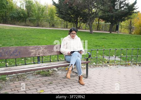 Jeune femme assise sur un banc dans le parc. Elle lit un livre tout en portant une veste douillette. Ses bottes et son jean s'accordent avec le cadre automnal. L'herbe verte et les arbres fr Banque D'Images
