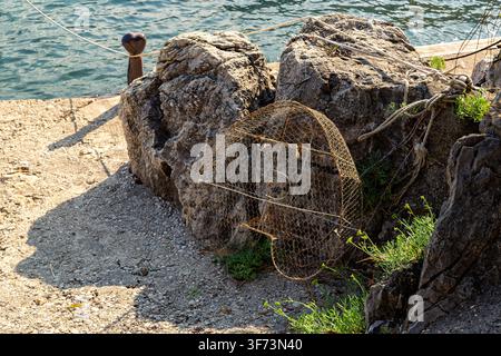 Un piège à crabes cylindrique repose contre de gros rochers sur une jetée à côté de l'eau calme de la mer Adriatique. Des cordes s'étendent vers le rivage près d'un poteau métallique. Banque D'Images