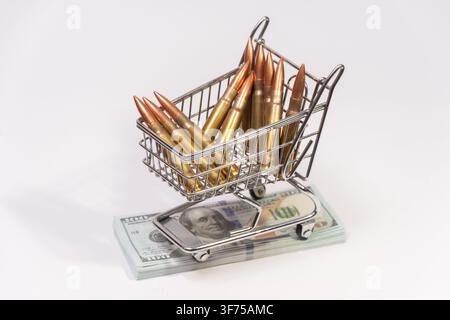 Un petit chariot de courses en métal chargé de cartouches de fusil en laiton est posé sur une pile de billets de cent dollars sur un fond blanc. Banque D'Images