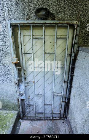 Grille de sécurité en métal rouillée couvrant une vieille porte en bois sur un mur en béton texturé, montrant la décrépitude et la négligence avec des toiles d'araignée et un luminaire cassé Banque D'Images