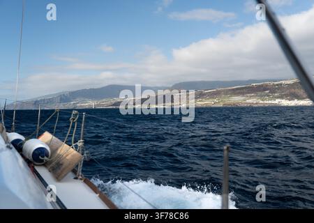 Un voilier de croisière navigue le long des côtes de La Palma, aux îles Canaries, sous un soleil éclatant de janvier, avec de l'eau blanche s'écoulant de la proue, l'île s'élevant au loin en terrasses cultivées et falaises rougeâtres jusqu'à une crête coiffée de nuages surmontée d'un phare. Banque D'Images