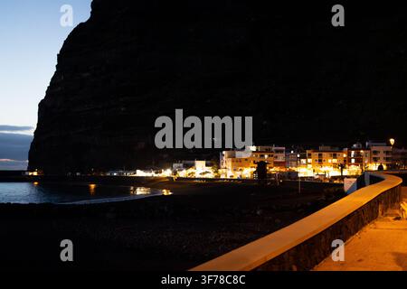 Le front de mer illuminé de Puerto de Tazacorte le long de sa plage de sable volcanique noir sur la côte ouest de La Palma, Îles Canaries, à l'heure bleue par une soirée claire de janvier, avec des bars et des immeubles d'habitation brillant sous une immense falaise sombre dominant la ville. Banque D'Images