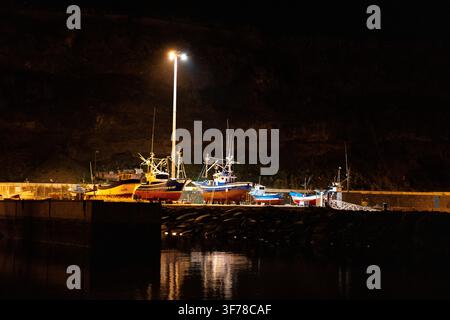 Bateaux de pêche en bois posés sur des berceaux sur la digue à Puerto de Tazacorte, La Palma, îles Canaries, éclairés par un seul projecteur du port contre la masse noire de la falaise en arrière-plan, leurs mâts se reflétant dans l'eau calme du port intérieur par une nuit claire de janvier. Banque D'Images